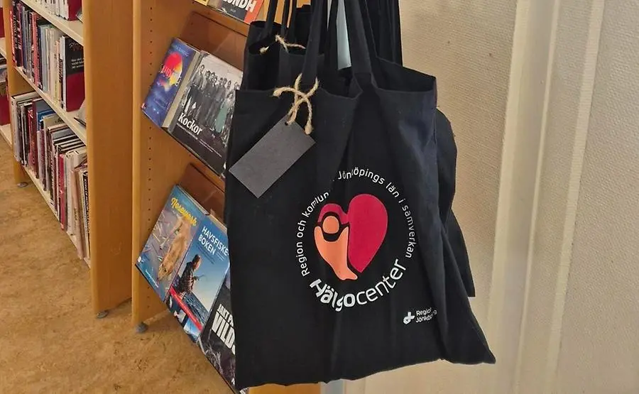 Svart tygpåse med logga Hälsocenter hänger i ett bibliotek.