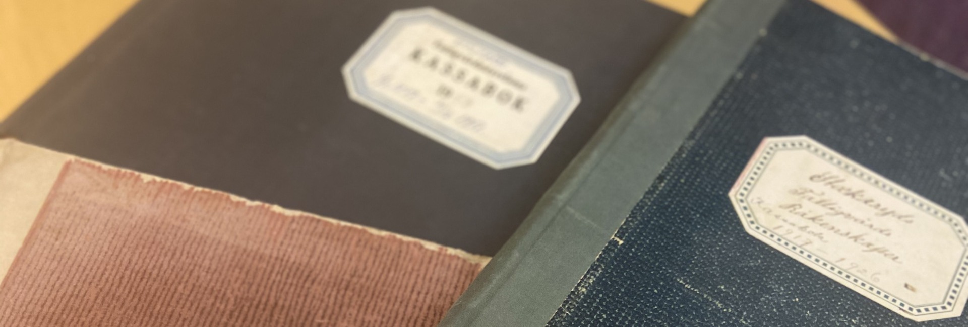 Tre gamla böcker i svart, blått och rött ligger på ett bord. Bilden är avskuren. Etiketterna är suddiga och därmed svåra att läsa ut. En bok är en gammal kassabok från 1940-talet.